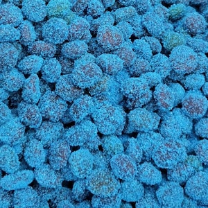 Puede incluir: Primer plano de un montón de caramelos azules. Los caramelos tienen un centro marrón oscuro y están cubiertos con un vibrante polvo azul azucarado. La textura parece áspera y granulada, lo que sugiere un dulce y posiblemente agrio.