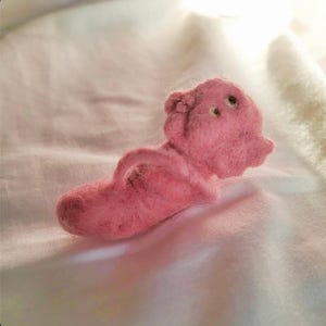 May include: A small, pink, needle-felted wool figure shaped like a stylized creature. The figure has a rounded head with two small, dark, circular eyes and a protruding, curved arm-like appendage extending from its textured, fuzzy body.