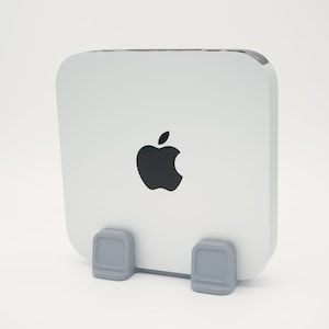 May include: A white Apple Mac Mini computer with the Apple logo in black. The computer is held by a grey stand. The computer has a rounded square shape.