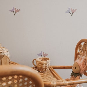 a white wall with pink and lilac flower decals. A small rattan table and chair with a baby doll.