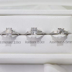 May include: Three moissanite engagement rings displayed in a white box. The rings are silver with different cut center stones: Emerald 2.0ct, Radiant 2.0ct, and Asscher 6.5mm. The brand name "AMELIA MINIMALIST moissanite jewelry" is at the top.