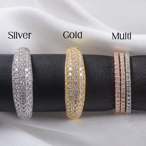 May include: Three rings, labeled "Silver", "Gold", and "Multi", are displayed on a black surface against a white backdrop. The silver and gold rings are each covered in small, clear stones. The multi ring set includes three thinner rings in rose gold, silver, and gold.
