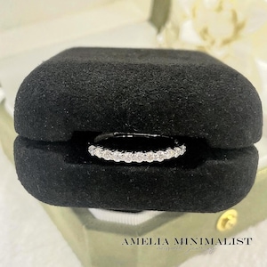 May include: A diamond eternity ring is presented in a black velvet ring box, set within a light green jewellery box. The ring showcases a line of small, round, white diamonds. The text "AMELIA MINIMALIST moissanite jewelry" is visible at the bottom.