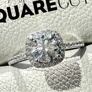 May include: A silver ring with a square-cut diamond surrounded by a halo of smaller diamonds. The ring is in a white box with the text "half eternity SQUARE CUT" on it.