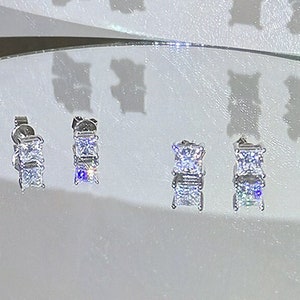 May include: Four pairs of silver earrings with clear square-shaped cubic zirconia stones. The earrings are arranged in a row on a white surface.