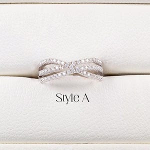 May include: A silver crossover band ring with small, clear gemstones. The ring is displayed in a white box with the text "Crossover Band" and "Style A". The brand name "AMELIA MINIMALIST" is also visible.