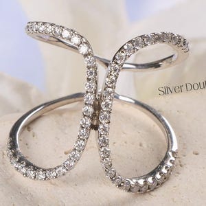 May include: A silver ring with a double C design, encrusted with small, sparkling stones. The ring is displayed on a light-colored, textured surface, with a blurred white flower in the background. The text "Silver Double C Ring" is visible.