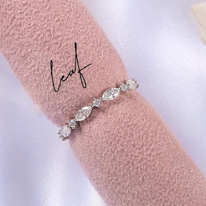 May include: A silver ring with alternating round and marquise-cut clear stones. The ring is displayed on a pink fabric with the word "leaf" written in cursive. The brand name "AMELIA MINIMALIST moissanite jewelry" is at the bottom.