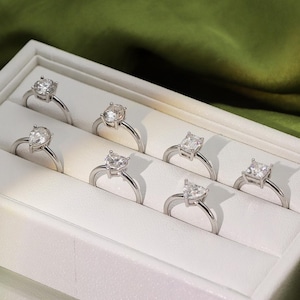 May include: A white box with a velvet interior displays a variety of silver rings with different shaped clear gemstones. The rings are arranged in rows on a white background. The text "Amelia Minimalist" is visible at the bottom of the image.