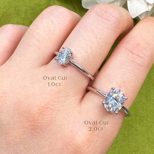 May include: Two oval cut diamond engagement rings on a hand. The top ring has a 1.0ct diamond and the bottom ring has a 2.0ct diamond. Both rings are set in a simple band.
