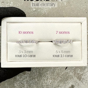 K&ouml;nnte beinhalten: Zwei Halb-Eternity-Ringe mit runden Brillanten. Der Ring links hat 10 Steine, jeder 3 mm im Durchmesser, insgesamt 1,0 Karat. Der Ring rechts hat 7 Steine, jeder 5 mm im Durchmesser, insgesamt 2,1 Karat. Die Ringe werden in einer wei&szlig;en Schachtel mit dem Text "ROUNDCUT half eternity" dar&uuml;ber pr&auml;sentiert.