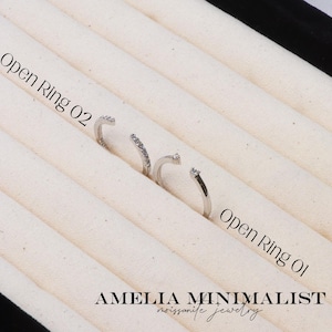 May include: Three open silver rings with clear gemstones are displayed on a cream-colored fabric. The rings are labeled "Open Ring 02" and "Open Ring 01". The brand name "AMELIA MINIMALIST moissanite jewelry" is at the bottom.