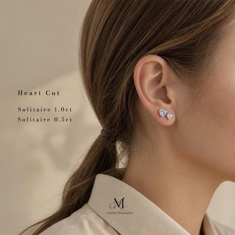 Puede incluir: Dos pendientes solitarios en forma de coraz&oacute;n con un engaste plateado. El texto en la imagen dice: Heart Cut, Solitaire 1.0ct, Solitaire 0.5ct y Amelia Minimalist. Los pendientes se llevan en el l&oacute;bulo de la oreja y tienen piedras facetadas.