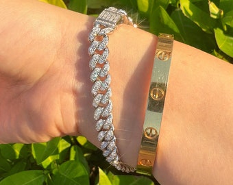 May include: A close-up of a woman's wrist wearing two bracelets. The bracelet on the bottom is a gold bangle with a repeating pattern. The bracelet on top is a silver chain bracelet with a clasp and is encrusted with small, clear stones.