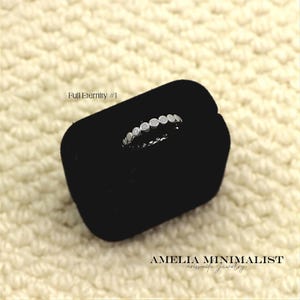 May include: A silver-toned full eternity ring featuring a series of small, round, bezel-set stones arranged in a continuous band. The ring is displayed on a black velvet holder. The text 'Full Eternity #1' and 'AMELIA MINIMALIST' are visible.