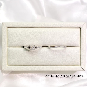 May include: Two silver rings displayed in a white jewelry box. The engagement ring features a square-cut center stone with smaller stones on the band. The wedding band is a delicate curved design with small stones. The text "AMELIA MINIMALIST moissanite jewelry" is at the bottom.