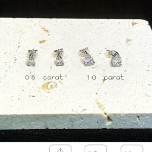 Puede incluir: Cuatro pendientes de bot&oacute;n plateados con corte de pera, dispuestos en dos pares. El primer par est&aacute; etiquetado como 0,5 quilates y el segundo como 1,0 quilates. El texto incluye amelia minimalist, CLASSIC Pear, Pear Cut, 0,5 Ct main stone y 1,0 Ct total stone.