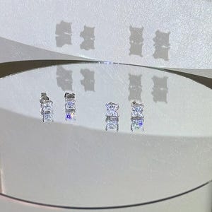 May include: Two pairs of silver-toned stud earrings featuring clear, square-cut stones in a four-prong setting. The earrings are displayed on a circular mirror surface, reflecting the light and casting shadows. The text 'amelia minimalist' is visible.