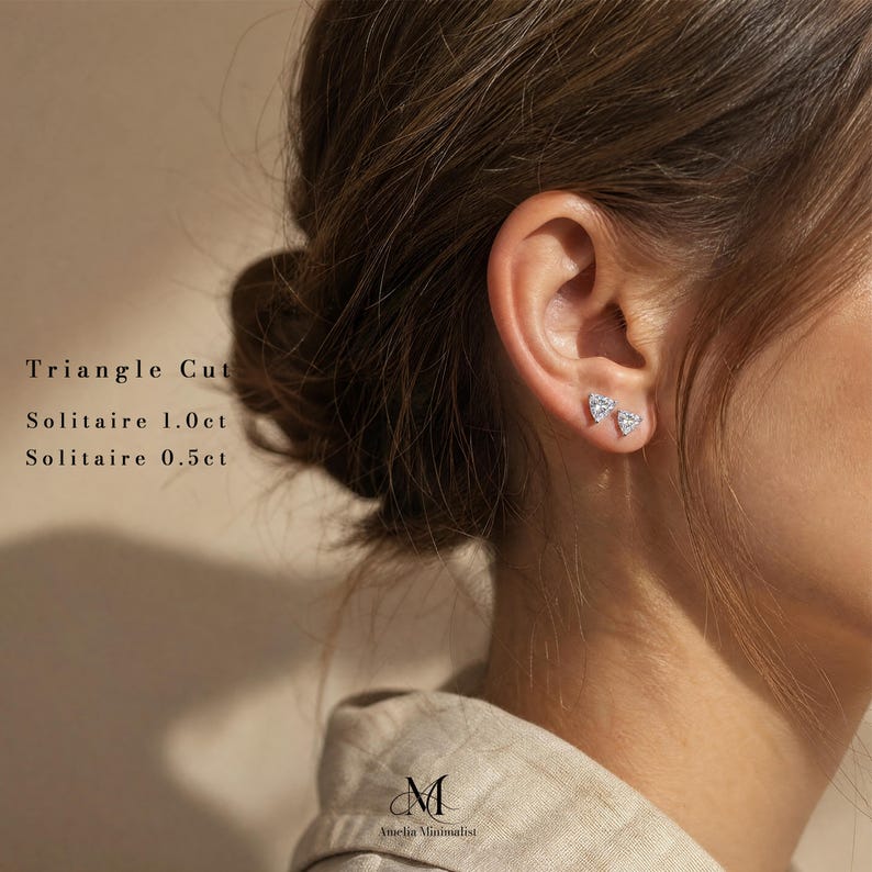 Puede incluir: Dos pendientes de bot&oacute;n plateados con piedras solitarias transparentes de corte triangular. El texto en la imagen dice: Triangle Cut, Solitaire 1.0ct, Solitaire 0.5ct, y el logotipo de Amelia Minimalist en la parte inferior central.