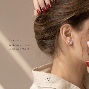 Puede incluir: Un par de pendientes de bot&oacute;n plateados con piedras solitarias en talla pera. El texto en la imagen dice: Pear Cut, Solitaire 1.0ct, Solitaire 0.5ct y el logotipo de Amelia Minimalist. Los pendientes se llevan en el l&oacute;bulo de la oreja.