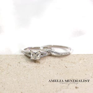 May include: Two silver rings, one with a square-cut center stone and a split band, the other with a thin band of small stones. The rings are on a light-colored surface. The text "AMELIA MINIMALIST" is visible.