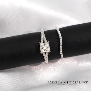 May include: Two silver rings. One ring features a square, clear gemstone at the center, with smaller stones set along the band. The second ring is a thin band with small, clear stones. The rings are displayed on a black surface against a white background.