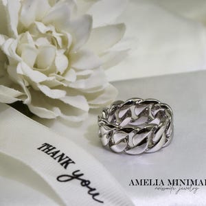 May include: A silver chain link ring sits on a white surface next to a white flower and a ribbon that says "THANK you". The ring is a thick band with a repeating chain design. The text "AMELIA MINIMALIST noissmile jewelry" is at the bottom.