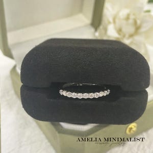 May include: A delicate ring with a row of small, sparkling white stones is displayed inside a black velvet ring box. The box is set within a light green jewelry box. The words "AMELIA MINIMALIST moissanite jewelry" are printed at the bottom.