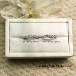 May include: Two half-eternity rings with clear stones are displayed in a white jewelry box. The rings are labeled "Half Eternity 9 stone" and "Half Eternity 10 stone." The brand name "AMELIA MINIMALIST moissanite jewelry" is at the bottom.