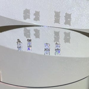 Puede incluir: Dos pares de pendientes de bot&oacute;n plateados con piedras transparentes de corte cuadrado en engastes de cuatro garras. Los pendientes se muestran sobre una superficie reflectante circular, proyectando sombras. El texto amelia minimalist es visible.