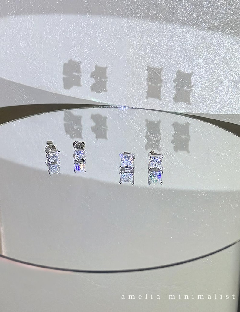 Puede incluir: Dos pares de pendientes de bot&oacute;n plateados con piedras transparentes de corte cuadrado en engastes de cuatro garras. Los pendientes se muestran sobre una superficie reflectante circular, proyectando sombras. El texto amelia minimalist es visible.
