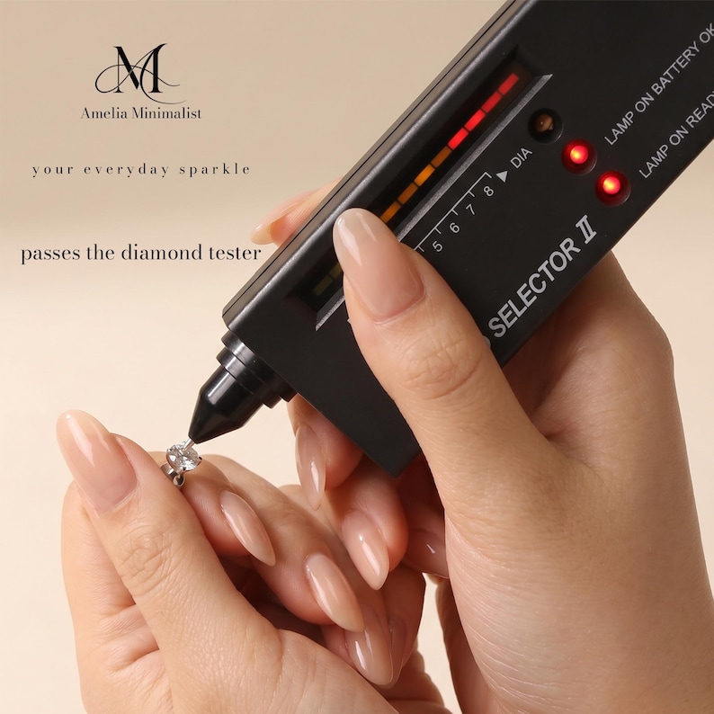 Puede incluir: Un dispositivo probador de diamantes electr&oacute;nico negro con una punta de sonda que toca una peque&ntilde;a piedra preciosa redonda y transparente. El dispositivo muestra una luz indicadora LED roja y el texto SELECTOR II. La imagen incluye el texto Amelia Minimalist, your everyday sparkle, y passes the diamond tester.