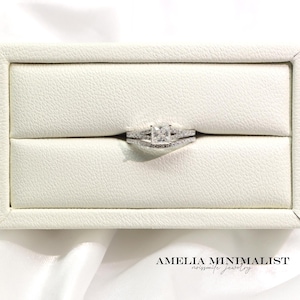 May include: A silver engagement ring set with a square-cut center stone and a delicate band, presented in a white jewelry box. The set includes a matching band with small accent stones. The text "AMELIA MINIMALIST moissanite jewelry" is visible.