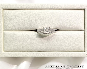 May include: A silver engagement ring set with a square-cut center stone and a delicate band, presented in a white jewelry box. The set includes a matching band with small accent stones. The text "AMELIA MINIMALIST moissanite jewelry" is visible.