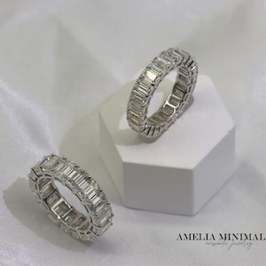 May include: Two silver eternity rings with emerald-cut stones. The rings are displayed on a white geometric surface against a white background. The rings are from Amelia Minimalist, moissanite jewelry.