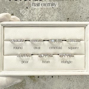 K&ouml;nnte beinhalten: Eine wei&szlig;e Pr&auml;sentationsbox mit sieben verschiedenen Stilen von Halb-Eternity-Ringen, die mit klaren Steinen besetzt sind. Die Ringe sind mit "rund", "oval", "Smaragd", "quadratisch", "Birne", "Herz" und "Dreieck" beschriftet. Der Text "7 STONES COLLECTION half eternity" befindet sich oben im Bild.