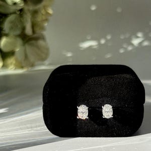 May include: A pair of oval-cut stud earrings with clear, faceted stones set in silver-toned metal. The earrings are displayed side-by-side on a black velvet jewelry box. The text AMELIA MINIMALIST is visible in the bottom right corner of the image.