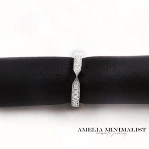 May include: A silver ring with a row of small, clear stones set along the top. The ring has a unique, twisted design. The ring is displayed on a black surface. The text "AMELIA MINIMALIST noissonite jewelry" is at the bottom.