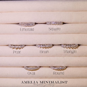 May include: Seven silver-toned rings with various gemstone cuts arranged in rows. The labels Emerald, Square, Pear, Heart, Triangle, Oval, and Round identify each style. The text AMELIA MINIMALIST moissanite jewelry is printed at the bottom.
