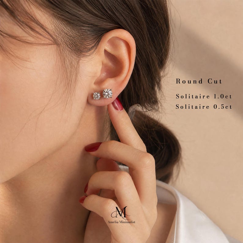 Puede incluir: Dos pendientes solitarios de corte redondo, uno de 1,0 ct y otro de 0,5 ct, con piedras transparentes facetadas engastadas en metal. El texto a la derecha dice Round Cut, Solitaire 1.0ct, Solitaire 0.5ct. Logo Amelia Minimalist abajo.