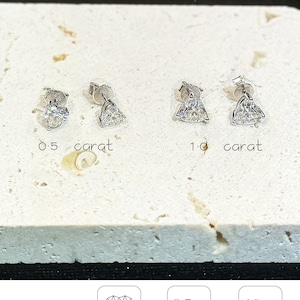 Puede incluir: Dos pares de pendientes de bot&oacute;n en forma de coraz&oacute;n, tono plateado, etiquetados como 0.5 quilates y 1.0 quilates. El texto incluye amelia minimalist, CLASSIC Heart, Heart Cut, 0.5 Ct each weight 5x5mm y 1.0 Ct each weight 6.5x6.5mm. Sobre una piedra texturizada.