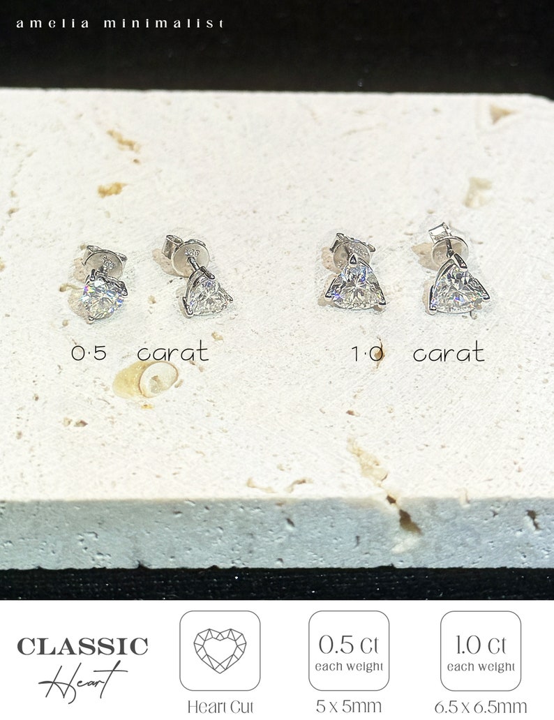 Puede incluir: Dos pares de pendientes de bot&oacute;n en forma de coraz&oacute;n, tono plateado, etiquetados como 0.5 quilates y 1.0 quilates. El texto incluye amelia minimalist, CLASSIC Heart, Heart Cut, 0.5 Ct each weight 5x5mm y 1.0 Ct each weight 6.5x6.5mm. Sobre una piedra texturizada.