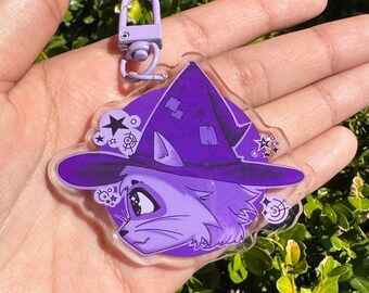 Puede incluir: Encanto de gato bruja morado y blanco con un sombrero morado y estrellas. El encanto está unido a un llavero morado.