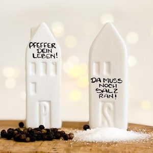 Könnte beinhalten: Weiße Salz- und Pfefferstreuer aus Keramik in Form von Häusern. Der Pfefferstreuer sagt "Pfeffer dein Leben!" und der Salzstreuer sagt "Da muss noch Salz ran!"