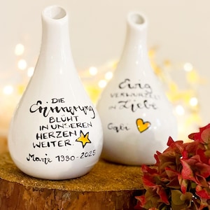 Könnte beinhalten: Zwei weiße Keramikvasen mit schwarzem handgeschriebenem Text und einem gelben Herz. Eine Vase hat einen Stern und den Text "Marie 1930-2025". Die Vasen stehen auf einem Holzsockel mit roten Blumen.