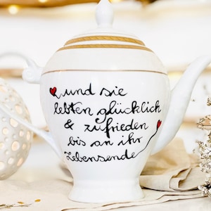 Könnte beinhalten: Weiße Teekanne mit goldener Verzierung und einer handgeschriebenen Botschaft in schwarzer Tinte, die lautet "und sie leben glücklich & zufrieden bis an ihr Lebensende".