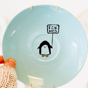 Könnte beinhalten: Hellblaue Keramikplatte mit einem Cartoon-Pinguin, der ein Schild mit der Aufschrift "FCK NZS" hält. Die Platte hat eine glänzende Oberfläche und goldfarbene Aufhängungen. Eine Person trägt einen beigen Strickpullover.