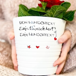 Könnte beinhalten: Weißer Keramik-Blumentopf mit roten Blumen und grünen Blättern. Der Topf hat den Text "Dein Wunschtext" dreimal darauf geschrieben. Außerdem sind drei rote Herzen und ein roter Stern auf den Topf gezeichnet.