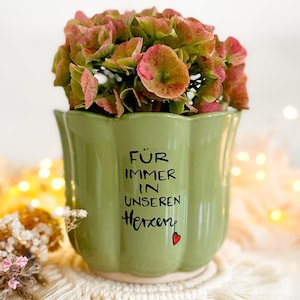 Könnte beinhalten: Ein grüner Keramik-Blumentopf mit gewelltem Rand, gefüllt mit rosa und grünen Blumen. Der Topf hat einen schwarzen Schriftzug "FÜR IMMER IN UNSEREN Herzen" mit einem kleinen roten Herz. Der Topf steht auf einem weißen Makramee-Untersetzer.