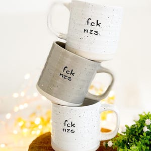 Könnte beinhalten: Drei Keramikbecher, gestapelt auf einem Holzsockel. Die Becher sind weiß und grau mit schwarzen Sprenkeln und dem Text "fck nzs". Jeder Becher hat einen Henkel.